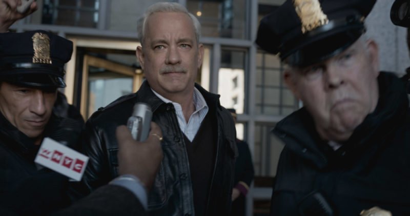 Tom Hanks als Sully wird interviewt und von Polizisten eskortiert in Sully