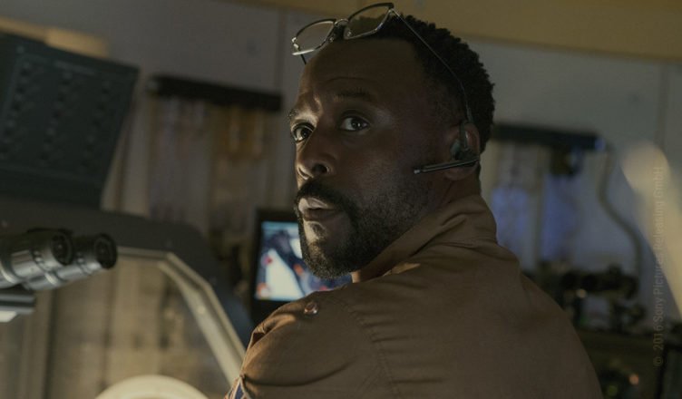 Hugh Derry (Ariyon Bakare) sitzt an seinem Arbeitsplatz und dreht sich überrascht um.
