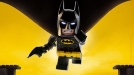 Die Lego Figur Batman fliegt vor einem gelben Hintergrund siegessicher der Kamera entgegen
