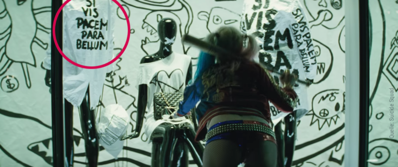 Harley Quinn klaut eine Handtasche aus einem eingeschlagenen Schaufenster in dem sich Kleidung mit dem Spruch Si vis Pacem Para Bellum befindet