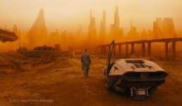 Blade Runner K steht neben einem Auto und starrt auf die rot-gelbe Skyline einer Mega City mit vielen Hochäusern.