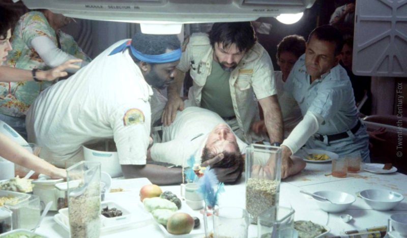 Die Crew in Alien 1 versammelt sich um einen Tisch auf dem ein infiziertes Besatzungsmitglied liegt