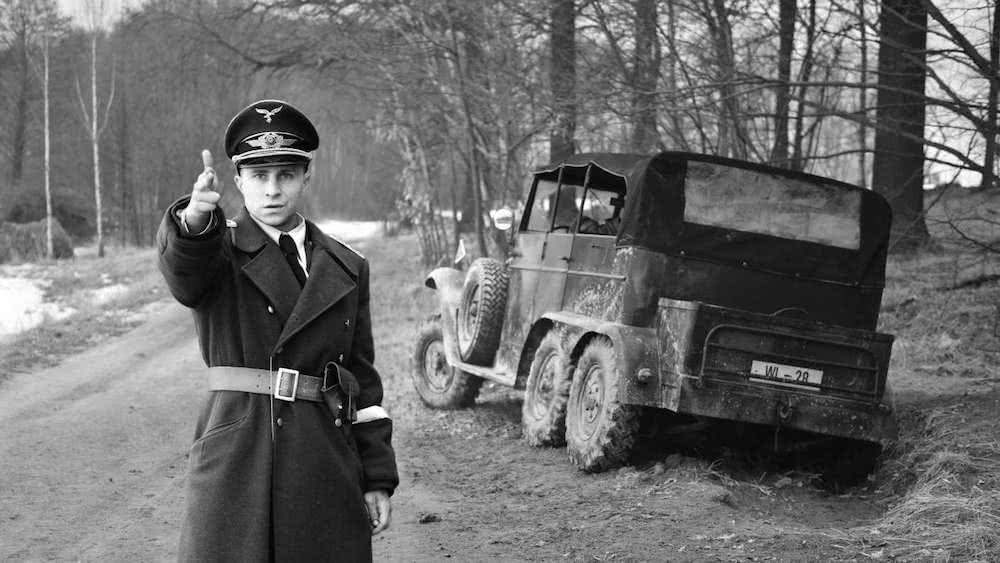 Max Hubacher als Willi Herold steht neben einem liegengebliebenen Geländewagen in einer SS-Uniform und zielt mit einer Fingerpistole in einem Szenenbild für Kritik Der Hauptmann