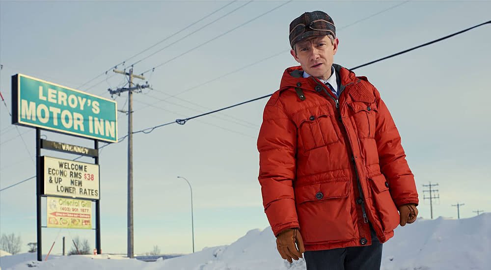 Martin Freeman in einer orangenen Daunenjacke vor dem verschneiten Schild eines Motels auf einem Plakat zu Fargo – Staffel 1