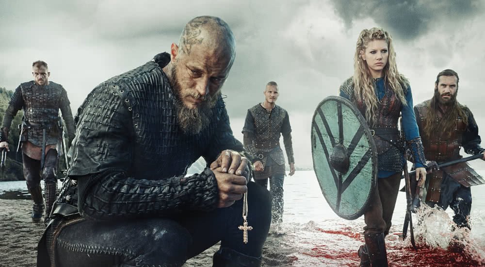 Travis Fimmel als Ragner Lothbrok und Katheryn Winnick als Lagertha sowie andere Hauptfiguren betreten schwer bewaffnet einen Strand der englischen Küste in Vikings Staffel 3