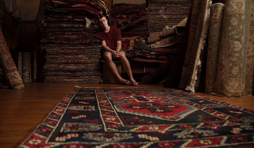 Ein Mann sitzt mit dem Kopf an einen Stapel Teppiche gelehnt und schaut auf einen türkischen Teppich, der vor ihm auf dem Boden liegt.